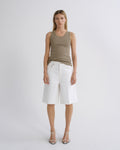 A model stands against a light gray studio background wearing a sleeveless taupe-brown tank top, white wide-leg shorts, and strappy high-heeled sandals.