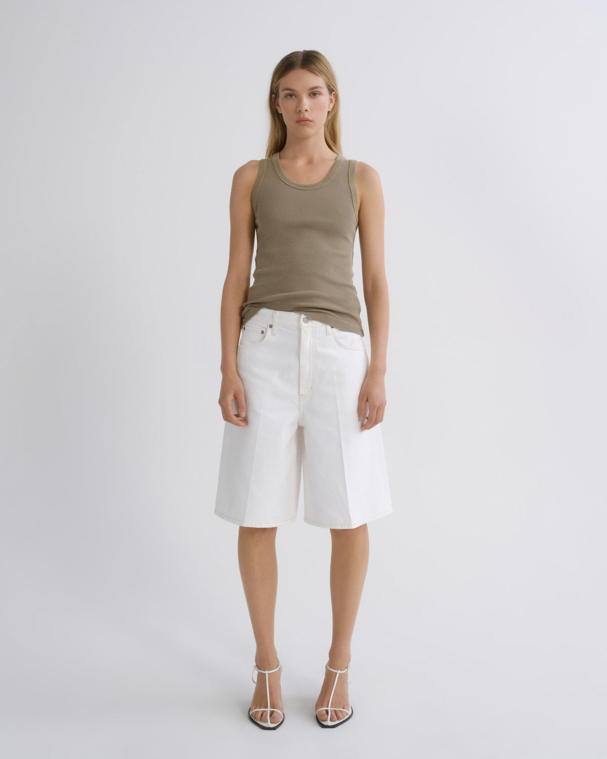 A model stands against a light gray studio background wearing a sleeveless taupe-brown tank top, white wide-leg shorts, and strappy high-heeled sandals.