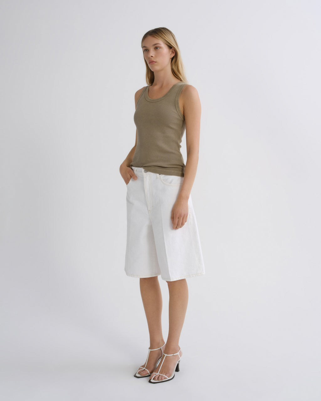 A woman modeling a sleeveless olive green tank top and white wrap-style shorts against a plain light gray studio background.