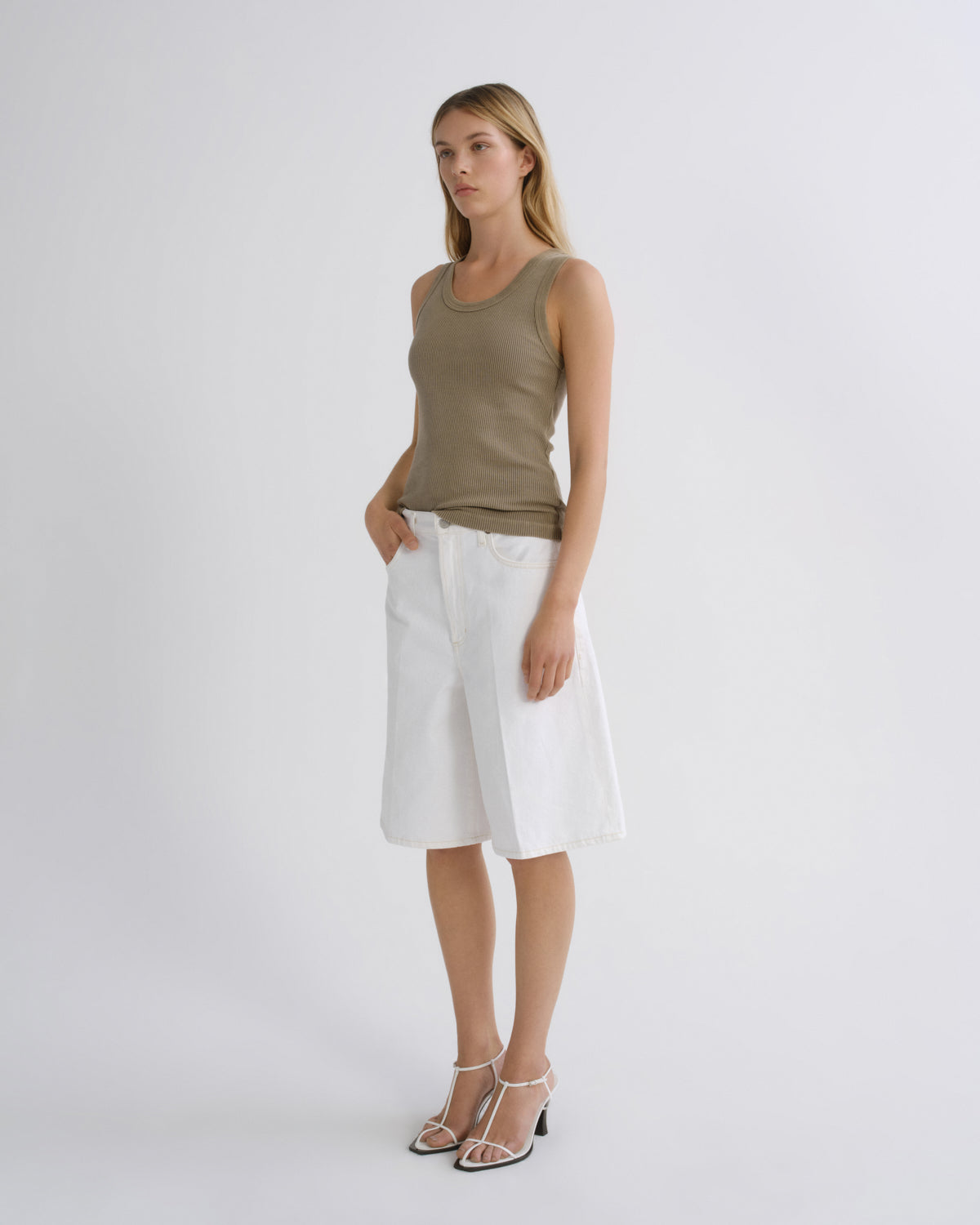 A woman modeling a sleeveless olive green tank top and white wrap-style shorts against a plain light gray studio background.