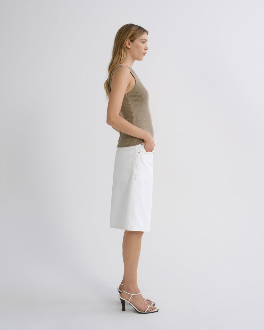 A woman stands in profile wearing a sleeveless taupe top and a white knee-length skirt with front pockets, paired with white strappy high-heel sandals, against a plain light gray studio background.