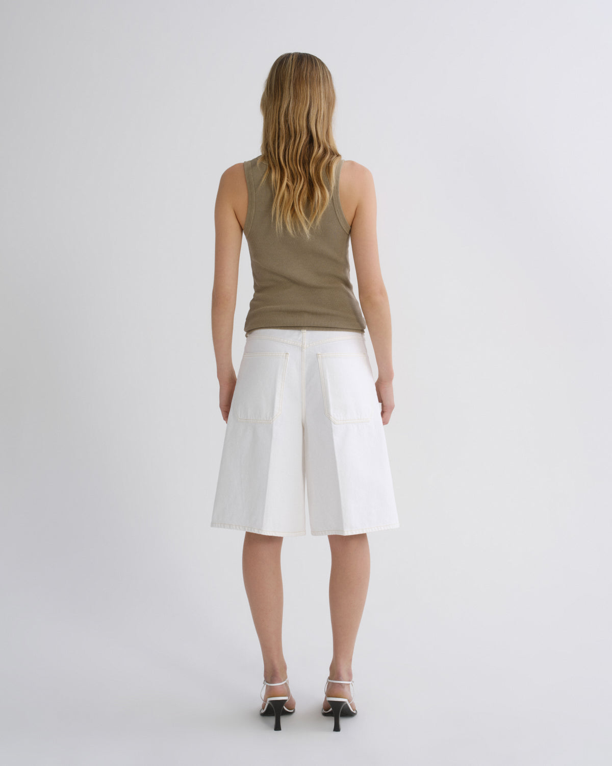 A woman viewed from behind wearing a sleeveless olive-brown top, white pleated shorts, and black high heels against a light gray studio background.