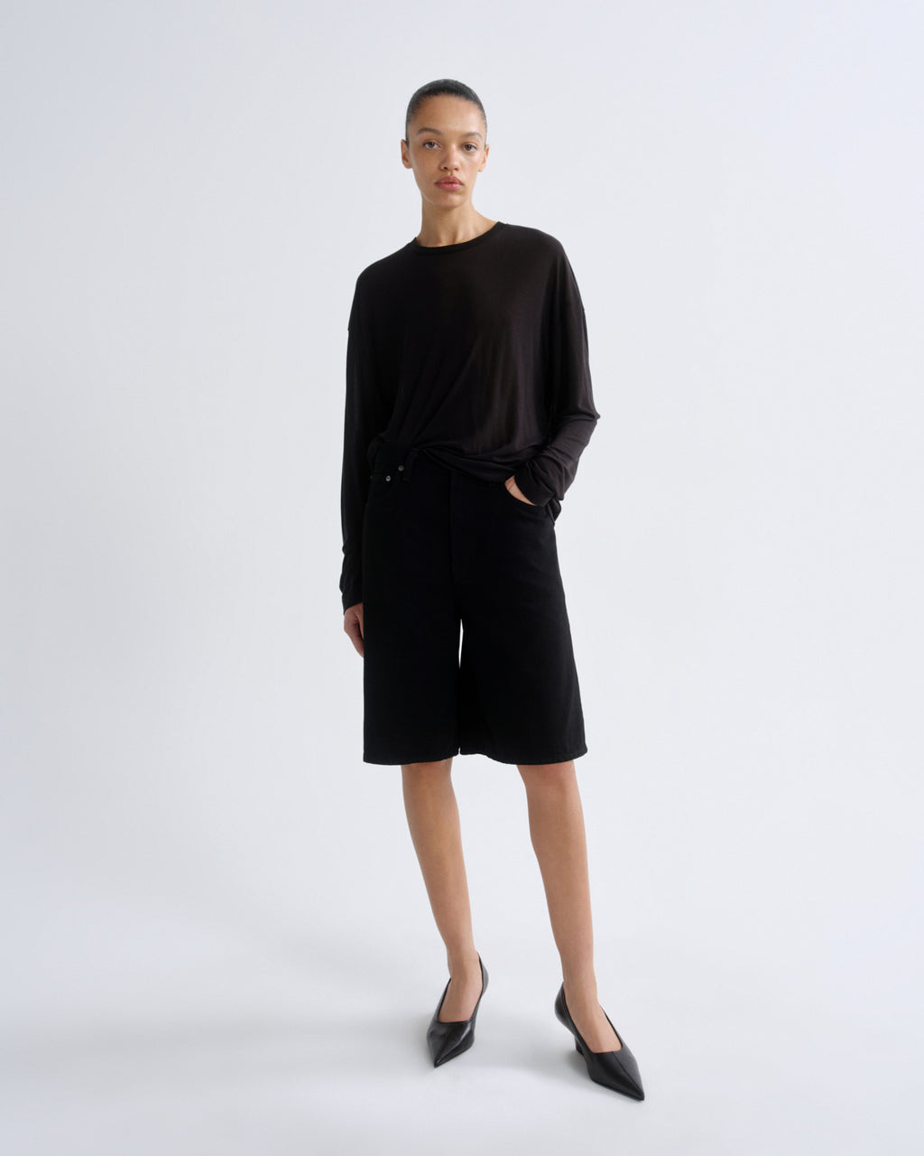 A model stands against a light gray studio background wearing a black long-sleeve top and matching black shorts, paired with black pointed flats.