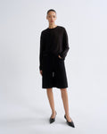 A model stands against a light gray studio background wearing a black long-sleeve top and matching black shorts, paired with black pointed flats.