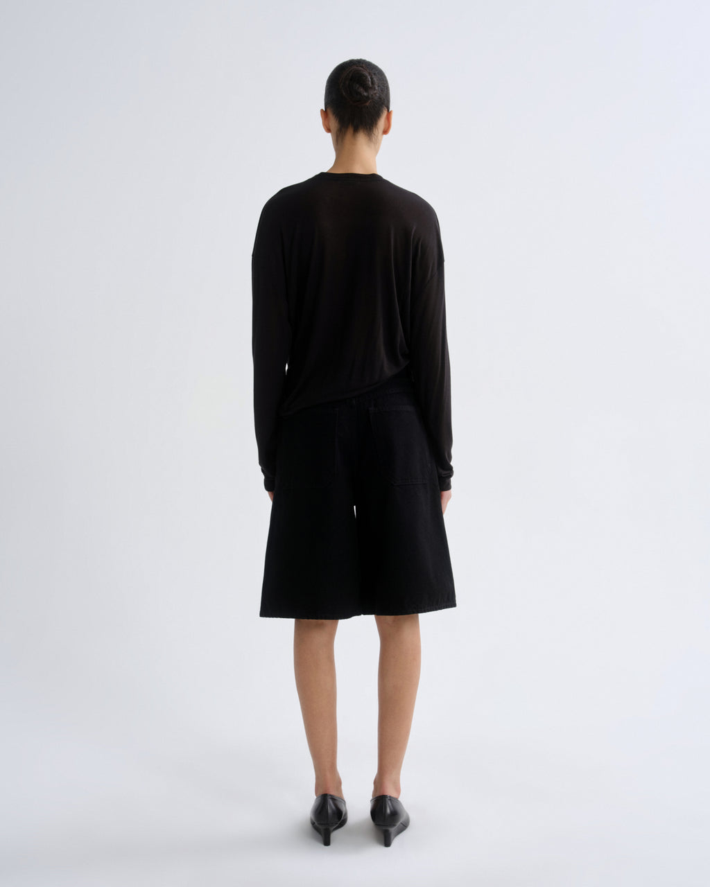 A model viewed from the back wearing a black long-sleeve top and matching black shorts, paired with black pointed shoes, against a plain light gray studio background.