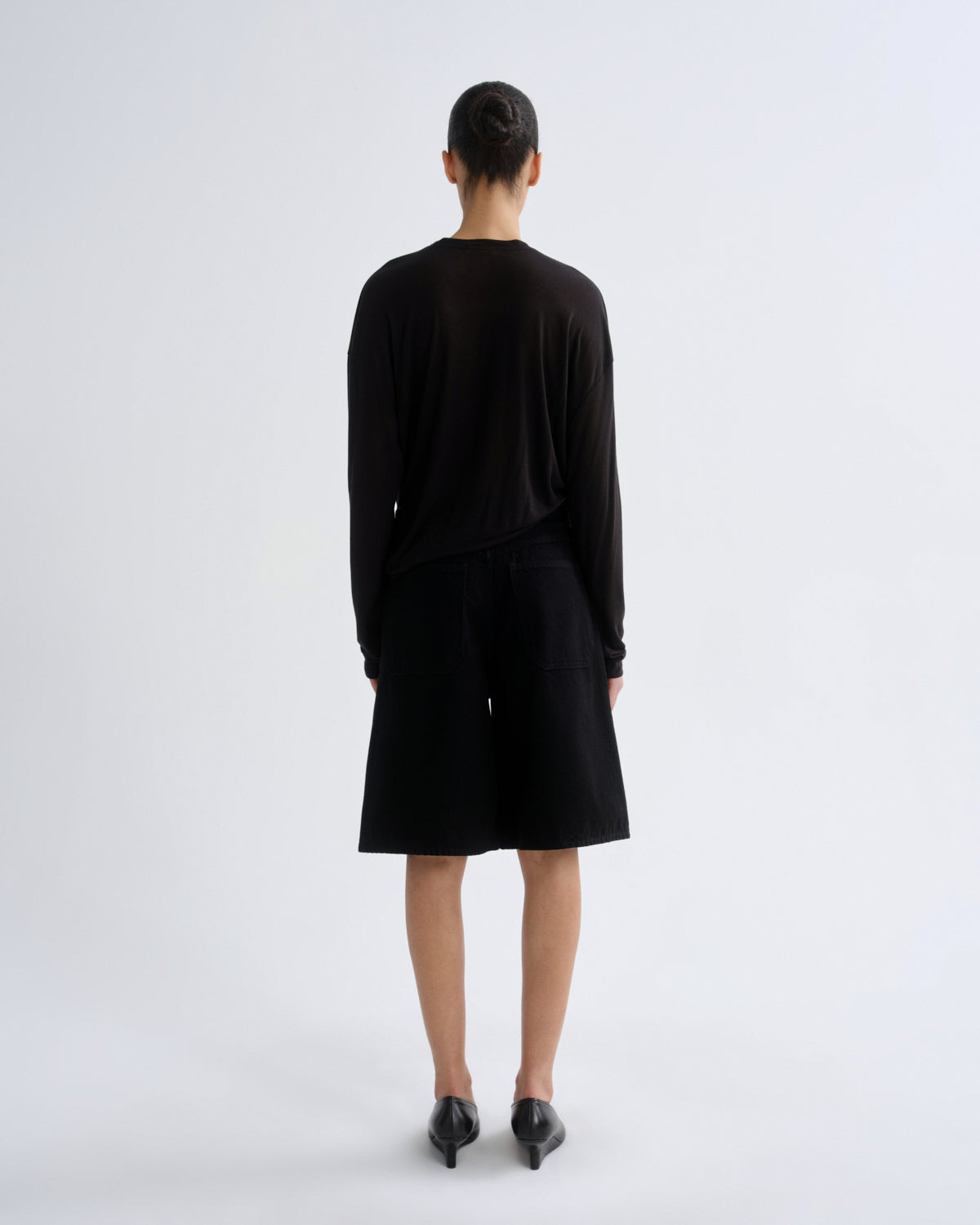 A model viewed from the back wearing a black long-sleeve top and matching black shorts, paired with black pointed shoes, against a plain light gray studio background.