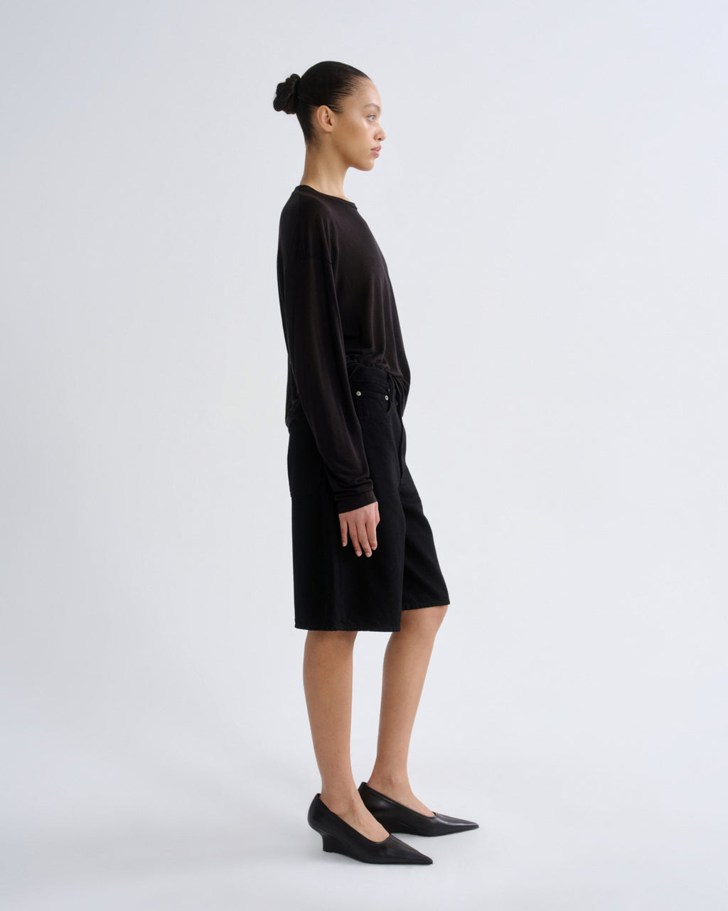 A model in side profile wearing a black long-sleeve top and matching black shorts, paired with black pointed-toe flats, against a light gray studio background.