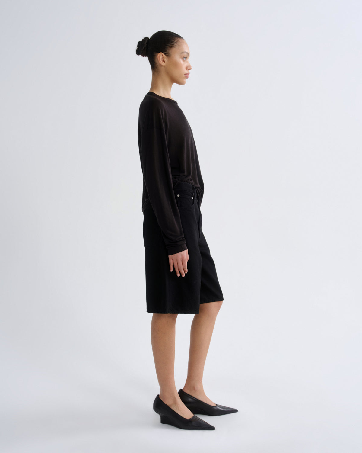 A model in side profile wearing a black long-sleeve top and matching black shorts, paired with black pointed-toe flats, against a light gray studio background.