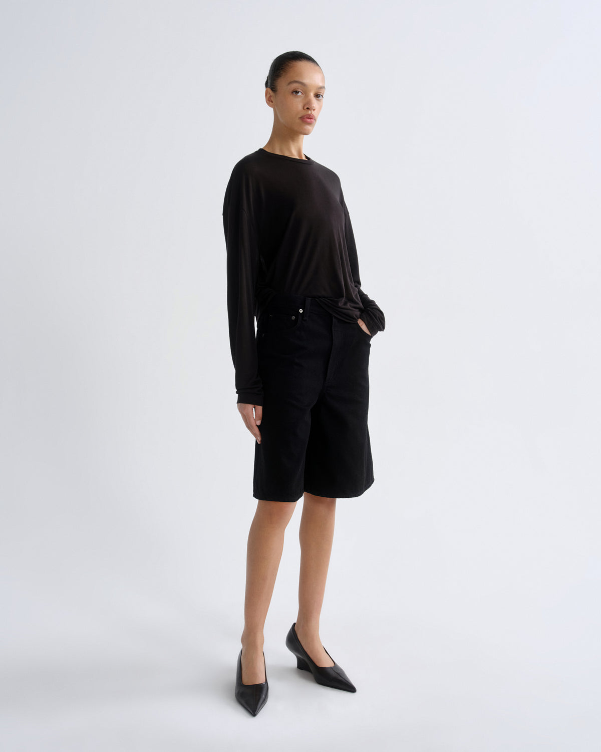 A model stands in a studio wearing a black long-sleeve top, black shorts, and black pointed-toe heels against a light gray background.