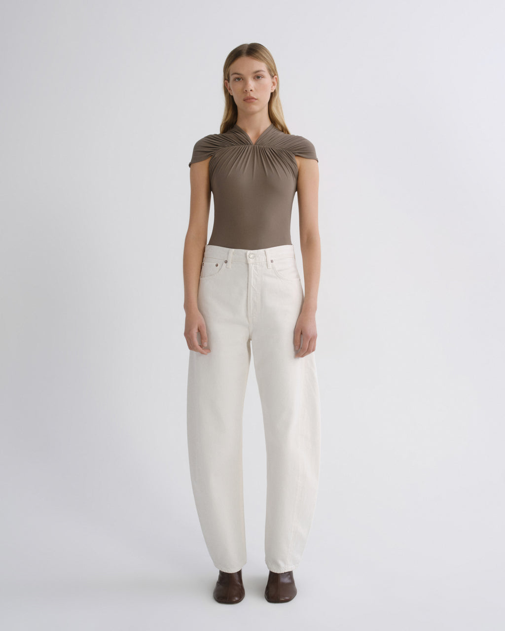 A model stands against a light gray studio background wearing a taupe-brown short-sleeve top with a gathered neckline and white wide-leg jeans.