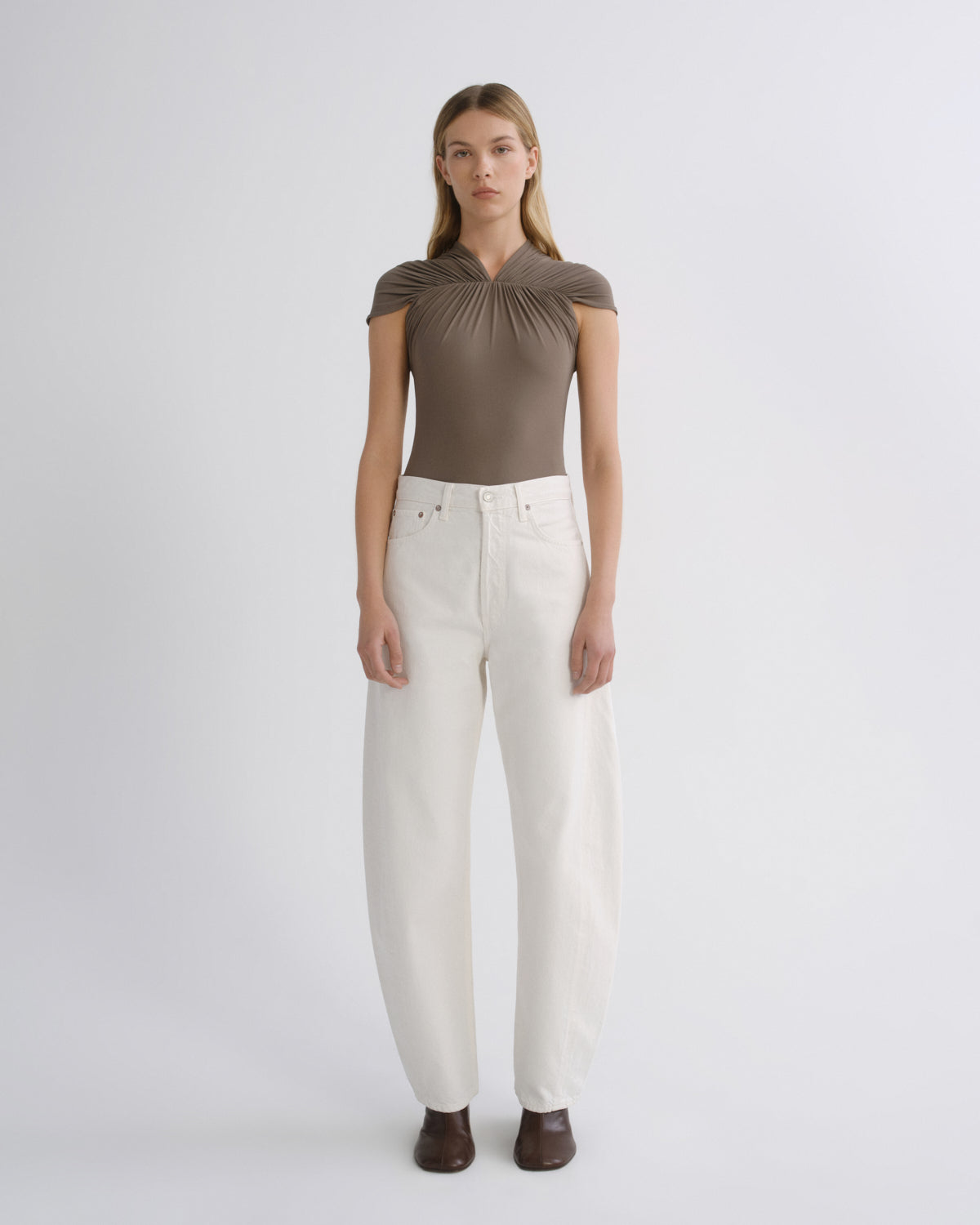 A model stands against a light gray studio background wearing a taupe-brown short-sleeve top with a gathered neckline and white wide-leg jeans.