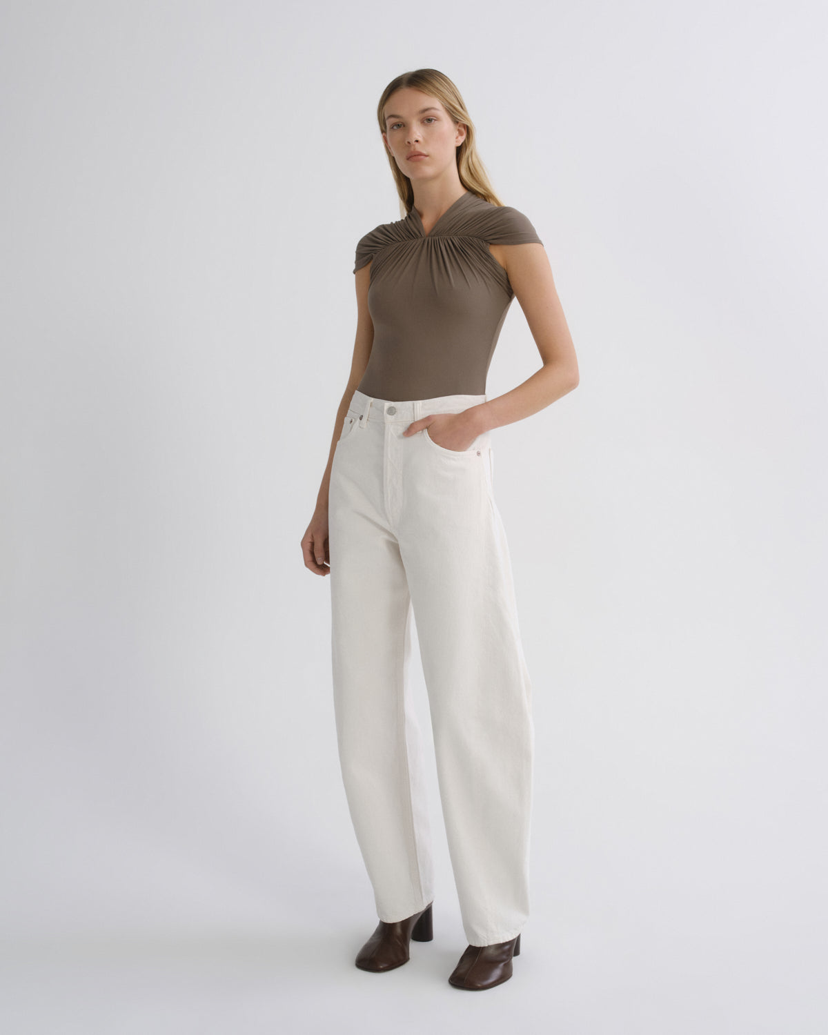 A model stands against a light gray studio background wearing a brown short-sleeve top with a gathered neckline, off-white wide-leg pants, and dark brown heeled shoes.