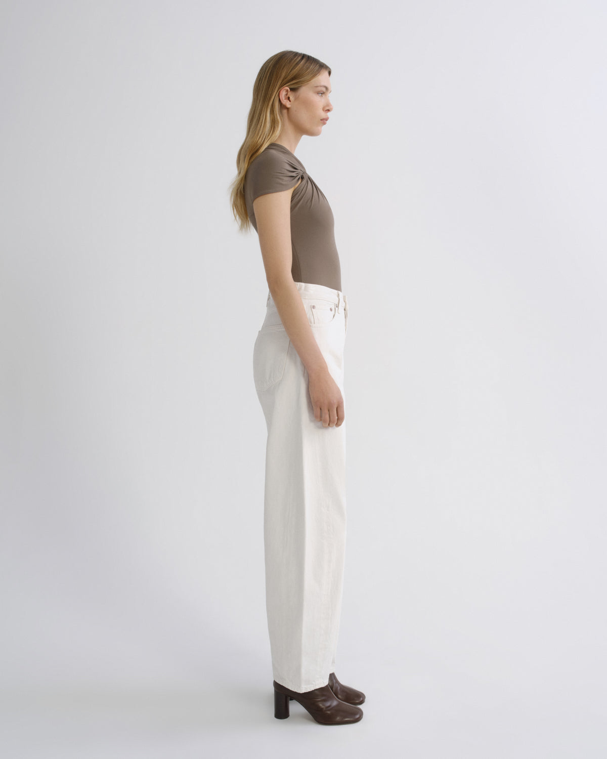A woman stands in profile wearing a taupe short-sleeve top, off-white wide-leg trousers, and dark brown heeled mules against a plain light gray studio background.