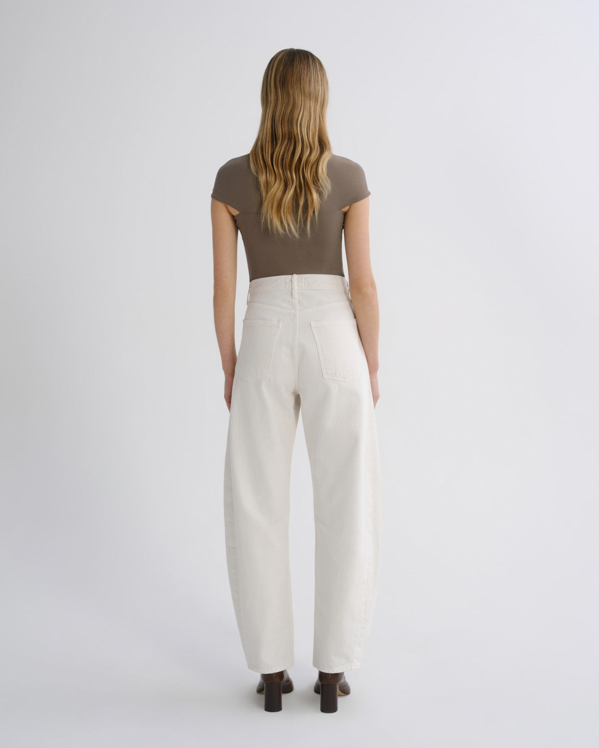 A woman viewed from behind wearing a brown short-sleeve top and off-white wide-leg trousers, standing against a plain light gray studio background.