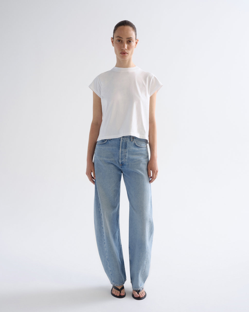 A model stands against a plain light gray studio background wearing a white short-sleeve crew-neck t-shirt and light-wash wide-leg blue jeans with black sandals.