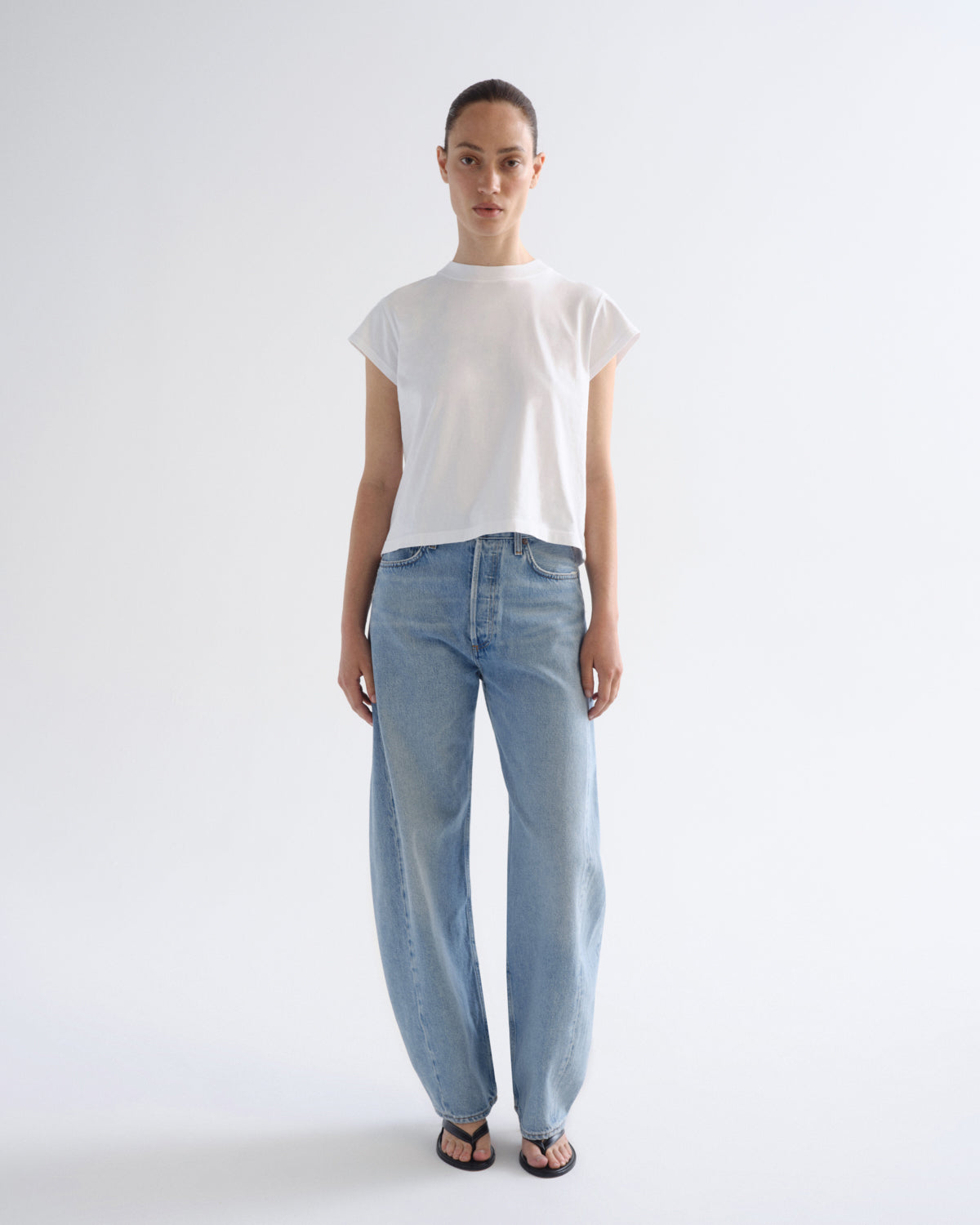 A model stands against a plain light gray studio background wearing a white short-sleeve crew-neck t-shirt and light-wash wide-leg blue jeans with black sandals.