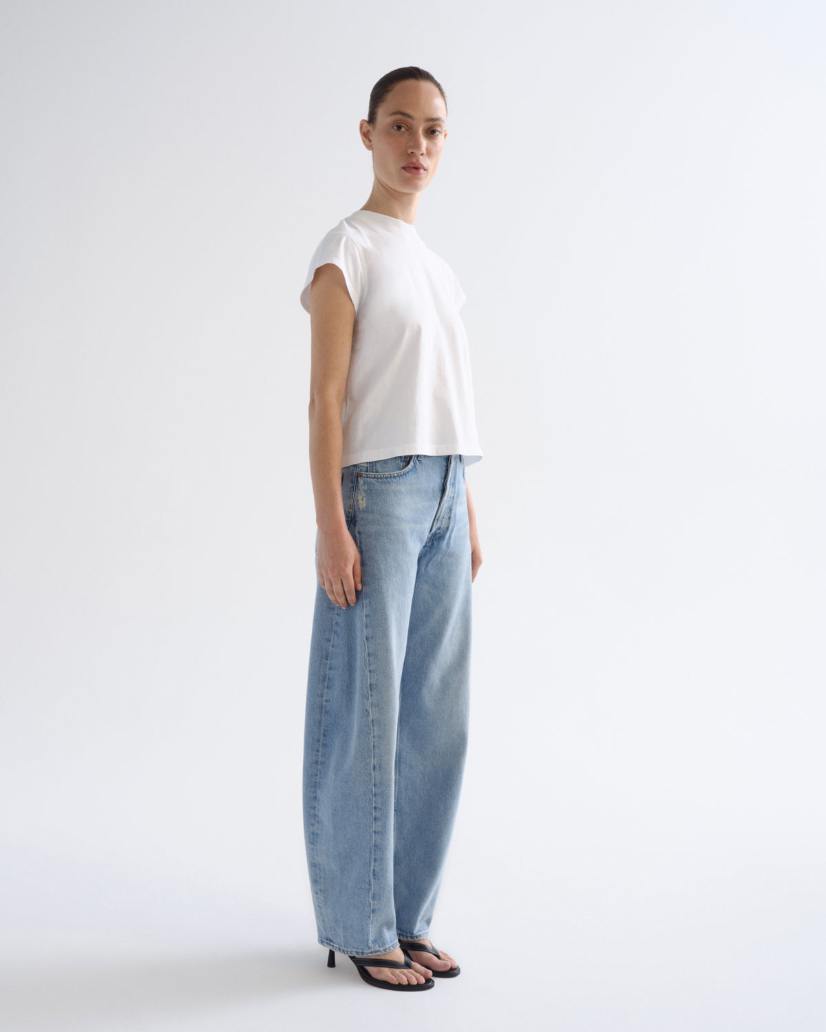 A model stands in profile wearing a white short-sleeve cropped T-shirt, light-wash wide-leg jeans, and black heeled sandals against a clean white studio background.