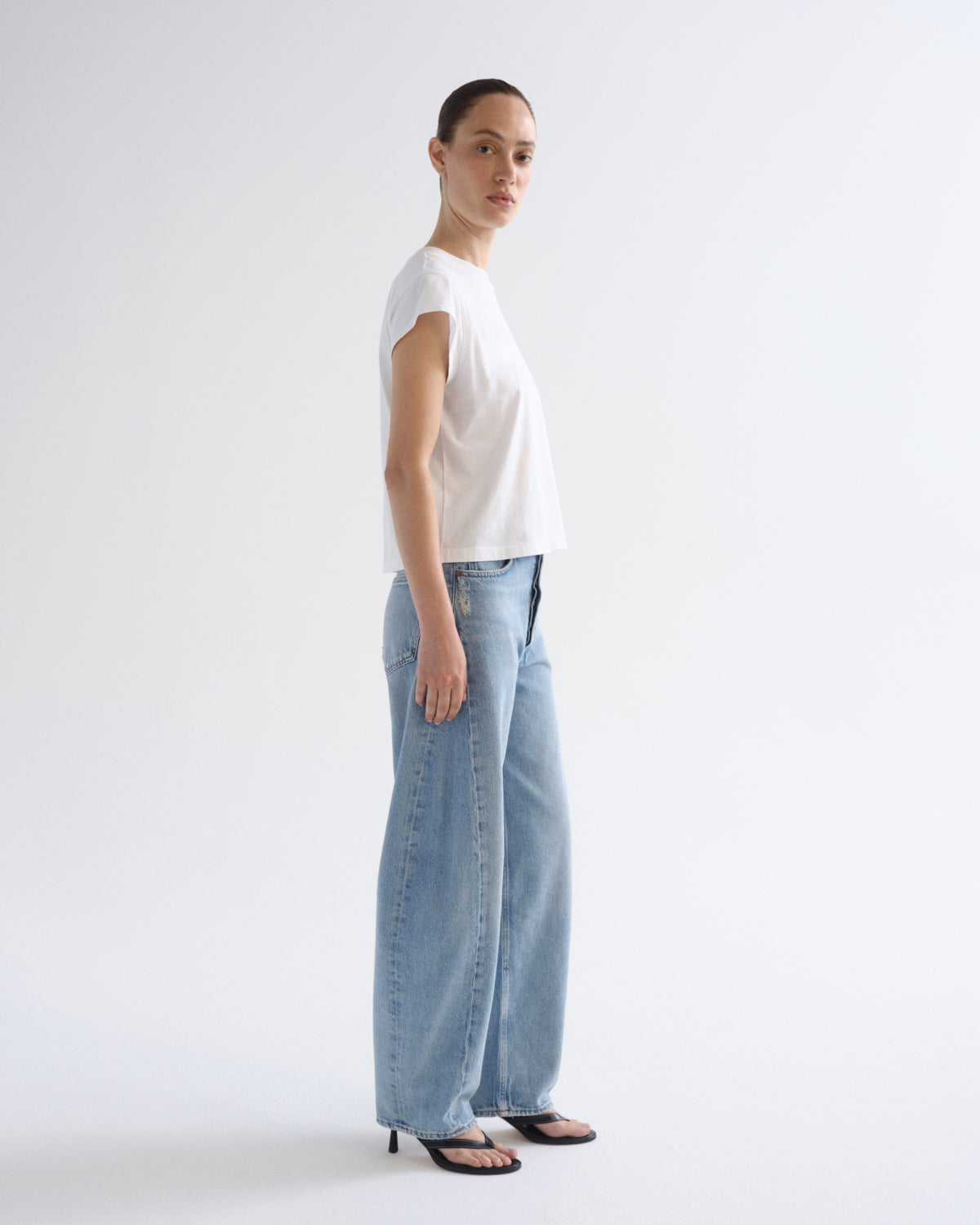 A woman stands in profile wearing a white short-sleeve crewneck t-shirt, light-wash wide-leg denim jeans, and black heeled sandals against a plain white studio background.