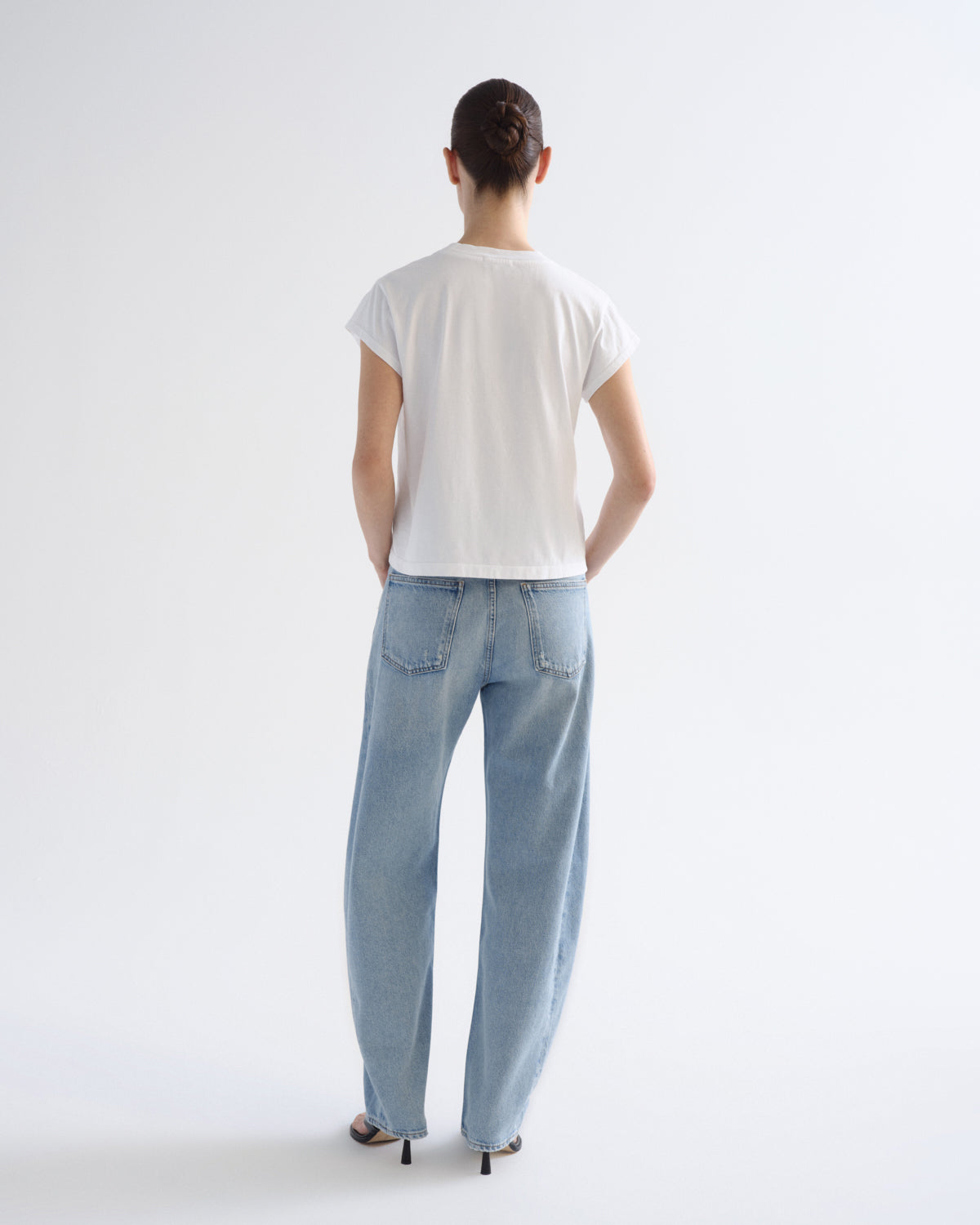 A model viewed from behind wearing a white short-sleeve t-shirt and light-wash wide-leg jeans, standing against a plain white studio background.