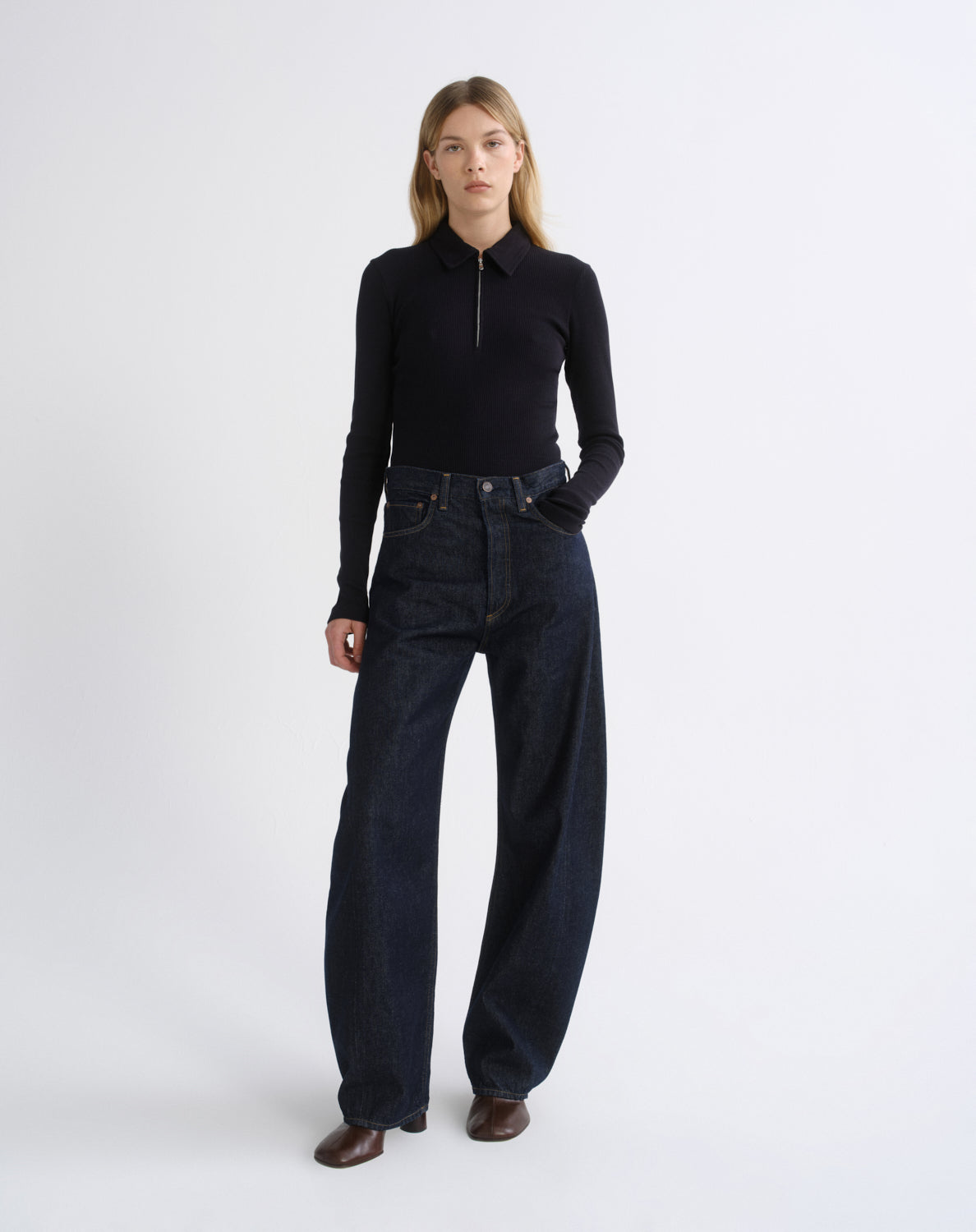 A model stands against a light gray studio background wearing a black long-sleeve half-zip mock-neck top and dark wide-leg denim jeans.