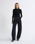 A model stands against a light gray studio background wearing a black long-sleeve half-zip mock-neck top and dark wide-leg denim jeans.