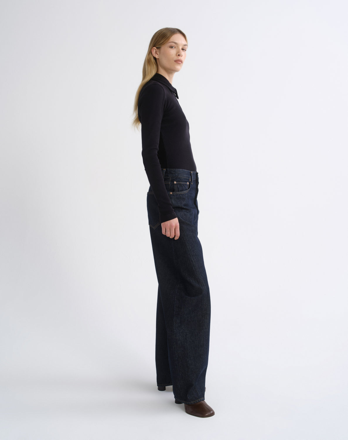 A woman stands in profile wearing a black long-sleeve top and dark wide-leg jeans against a clean light gray studio background.