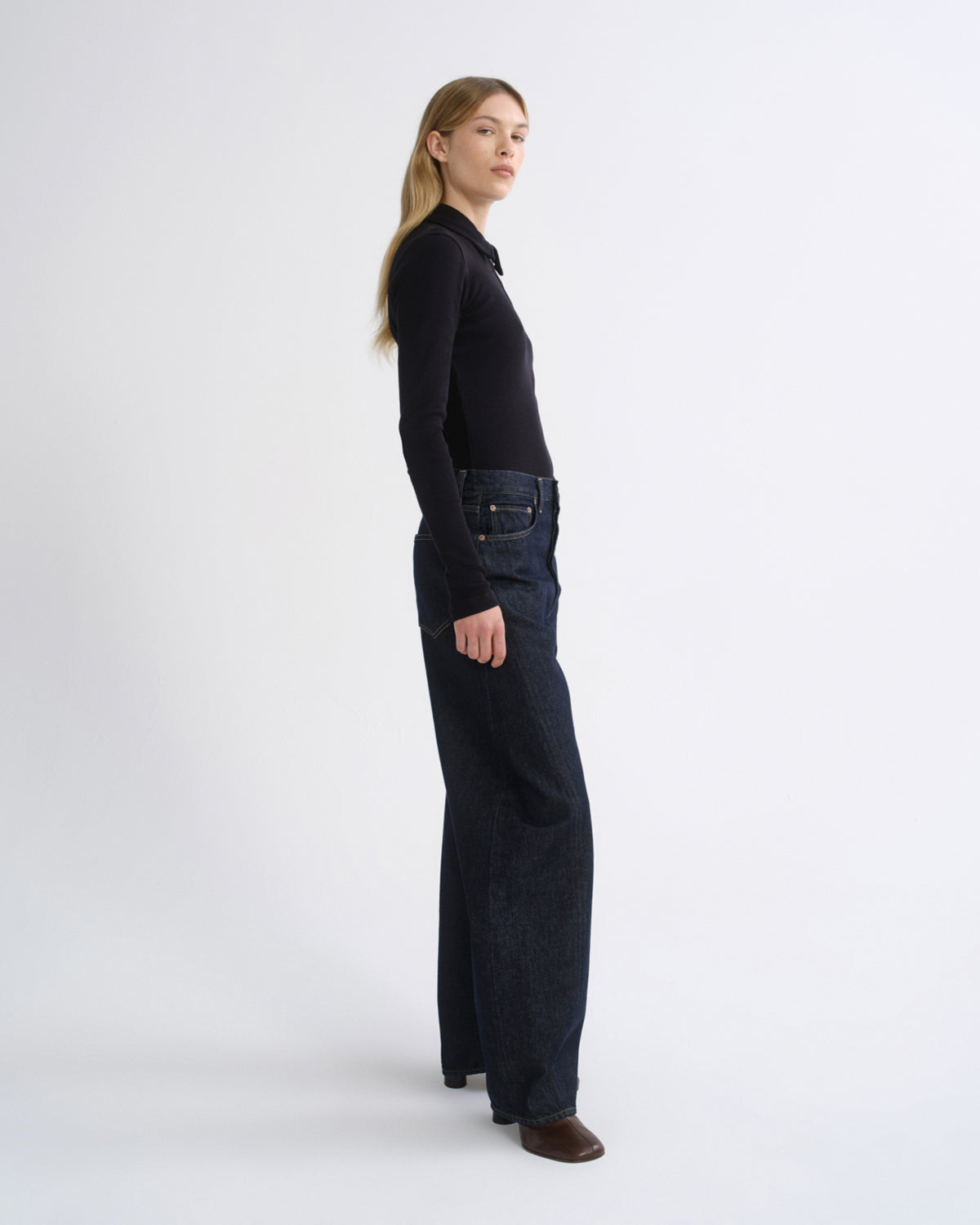 A woman stands in profile wearing a black long-sleeve top and dark wide-leg jeans against a clean light gray studio background.