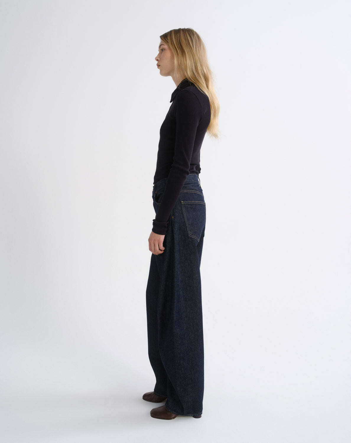 A woman stands in profile wearing a black long-sleeve top and dark wide-leg denim pants against a plain light gray studio background.