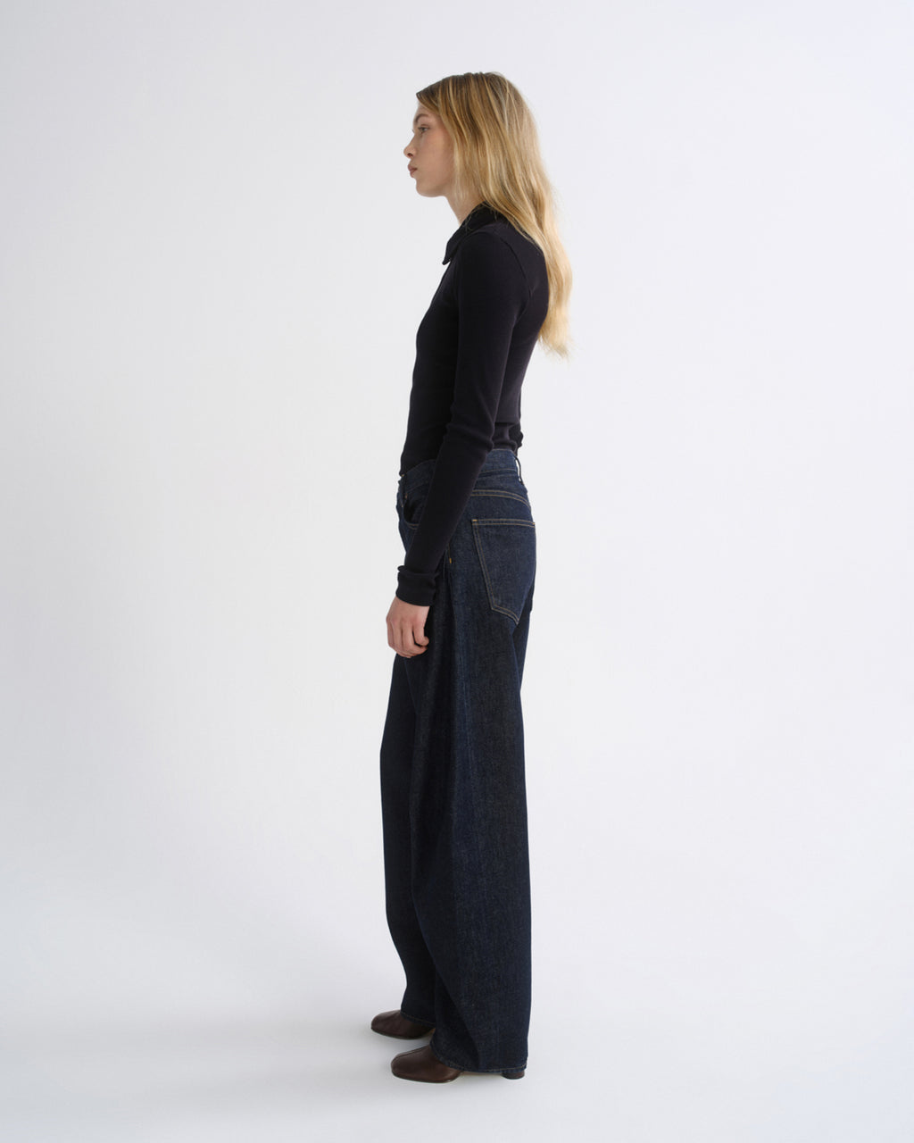 A woman stands in profile wearing a black long-sleeve top and dark wide-leg denim pants against a plain light gray studio background.