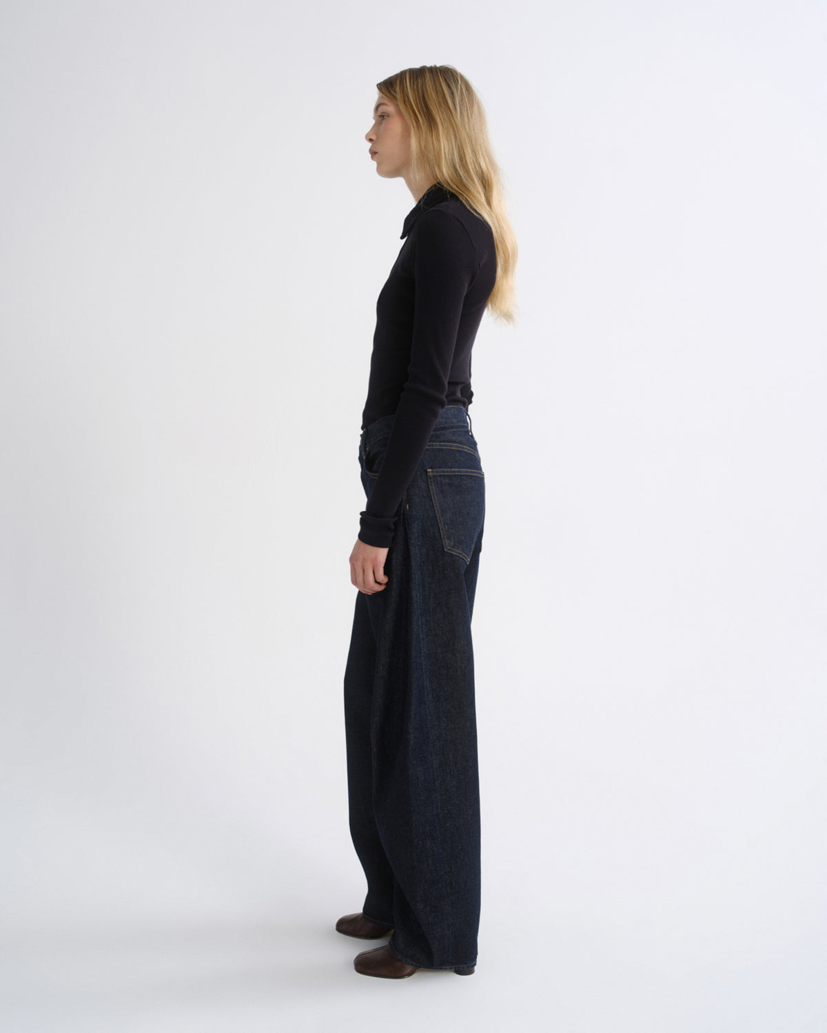 A woman stands in profile wearing a black long-sleeve top and dark wide-leg denim pants against a plain light gray studio background.