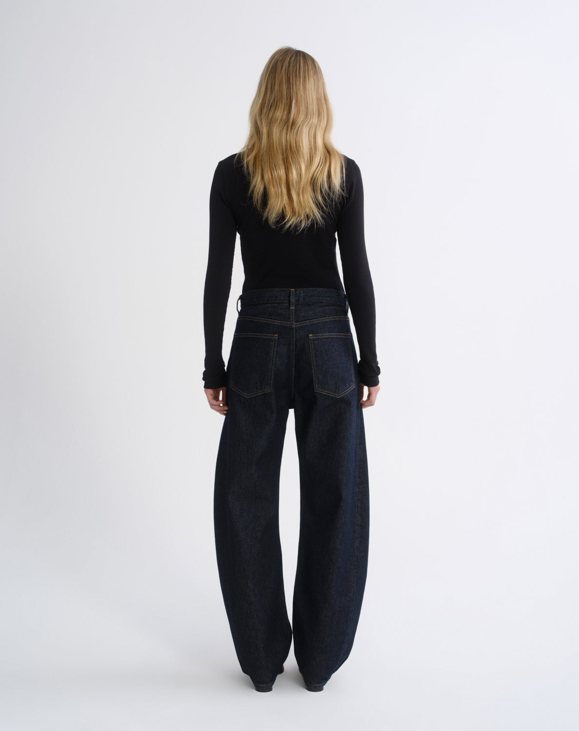 A woman viewed from behind wearing a black long-sleeve top and dark wide-leg denim pants against a plain light gray studio background.