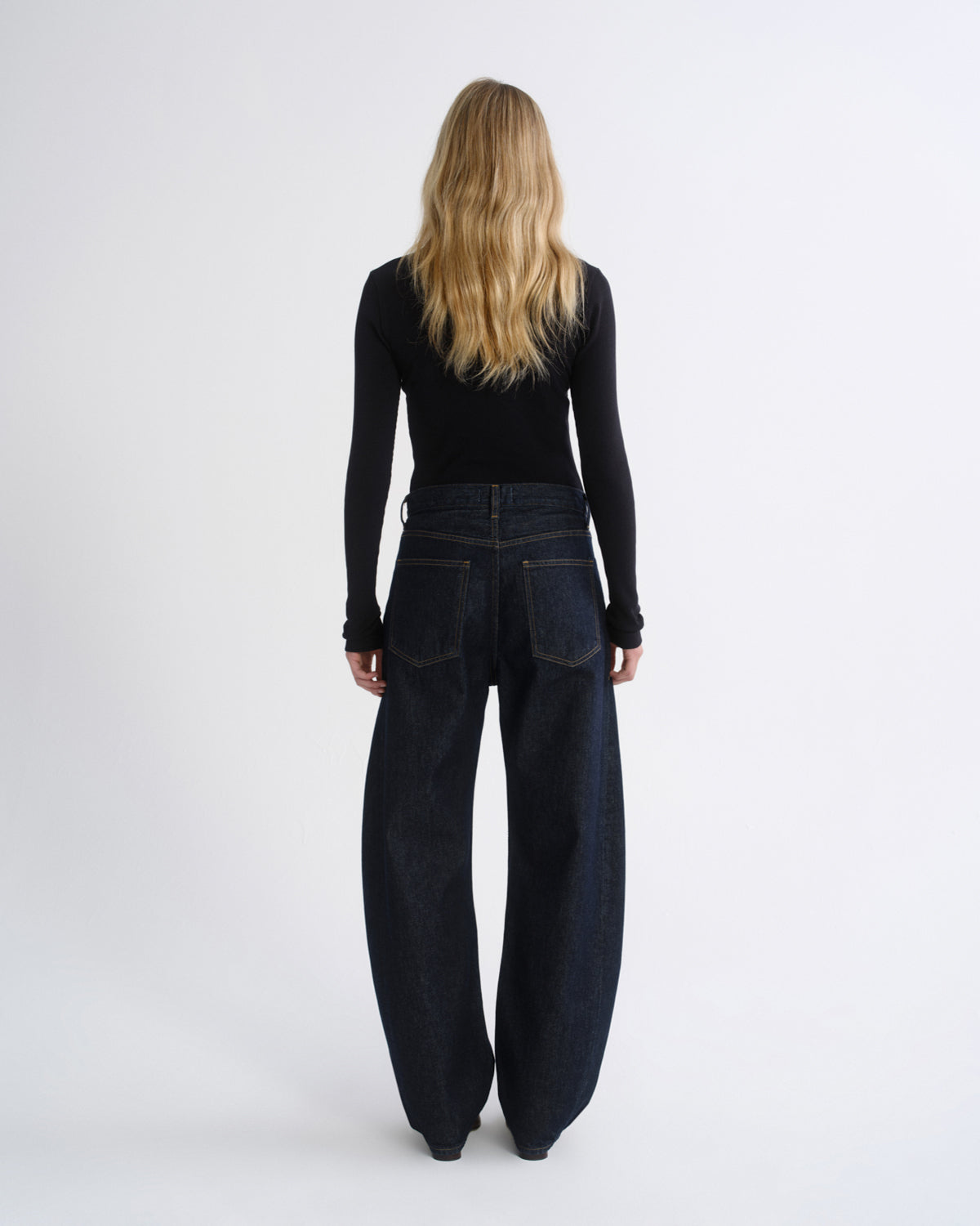 A woman viewed from behind wearing a black long-sleeve top and dark wide-leg denim pants against a plain light gray studio background.