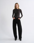 A model stands against a light gray studio background wearing a dark brown long-sleeve top with a ruched, crisscrossed texture and black wide-leg trousers.