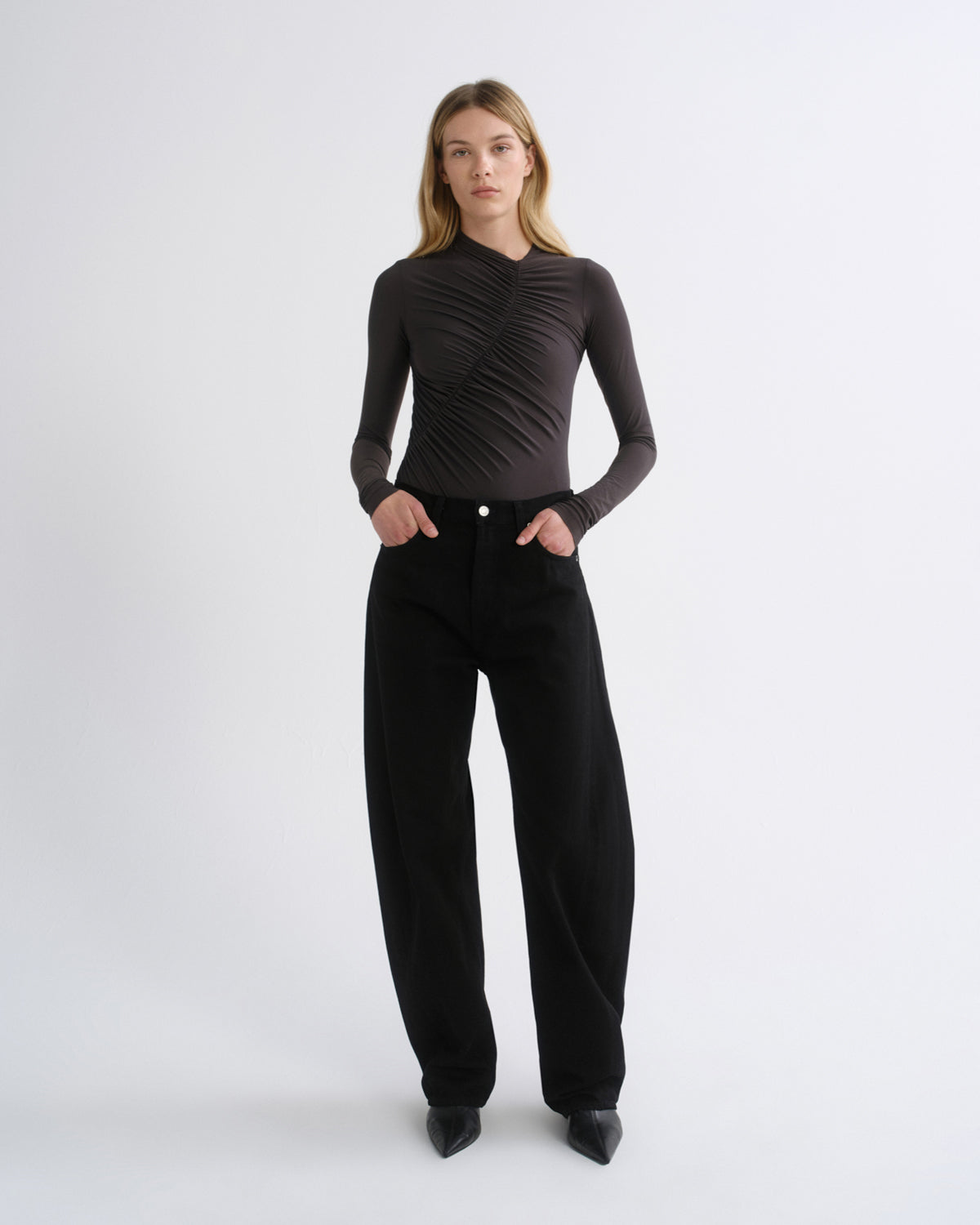 A model stands against a light gray studio background wearing a dark brown long-sleeve top with a ruched, crisscrossed texture and black wide-leg trousers.
