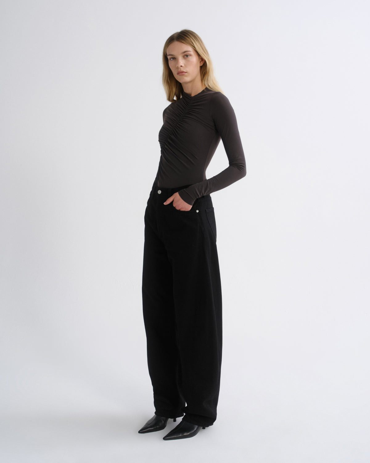 A woman modeling a black long-sleeve top with a ruched, gathered neckline and wide-leg black trousers, standing against a plain light gray studio background.
