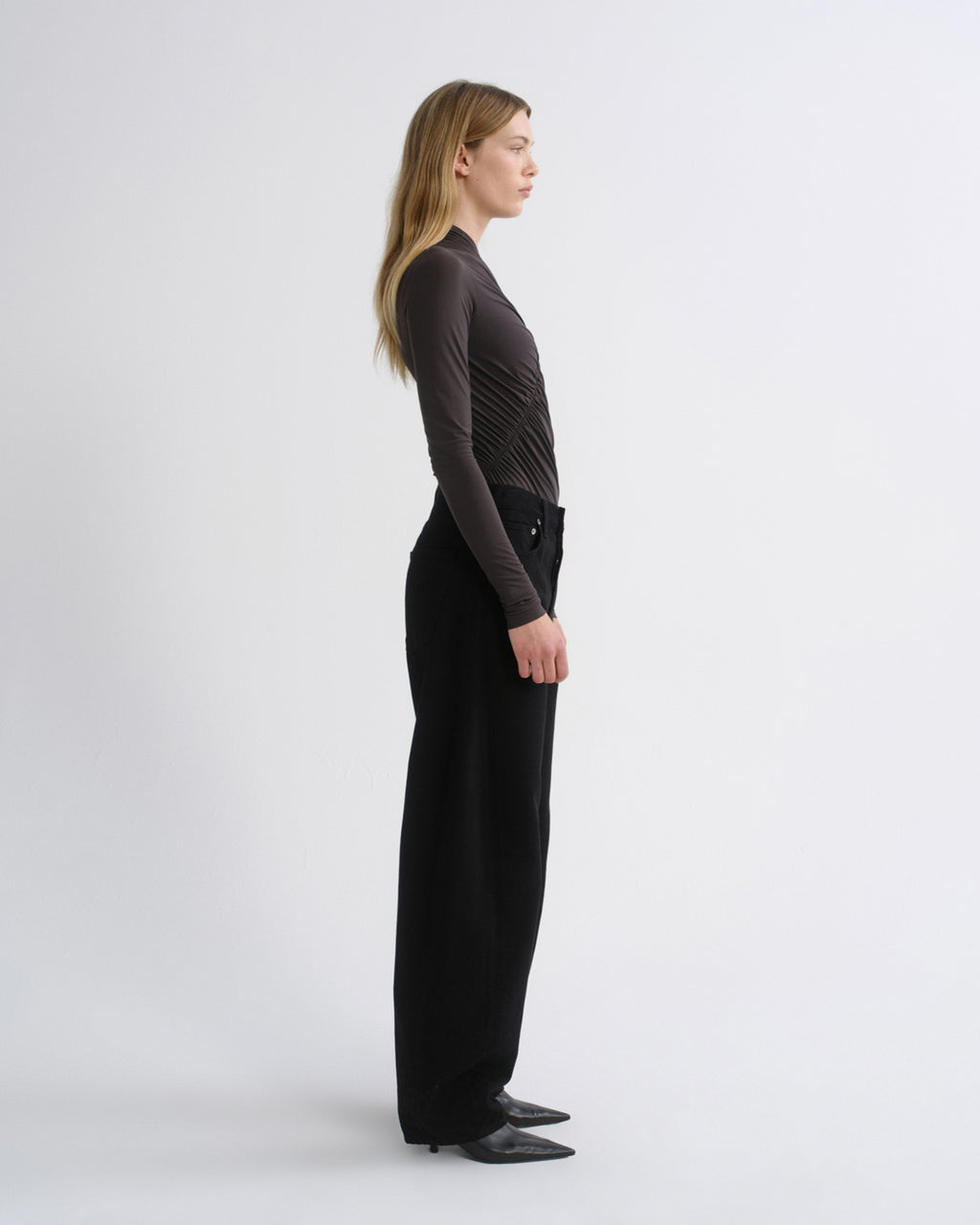 A woman stands in profile wearing a dark brown long-sleeve ribbed top and black wide-leg trousers against a plain light gray studio background.