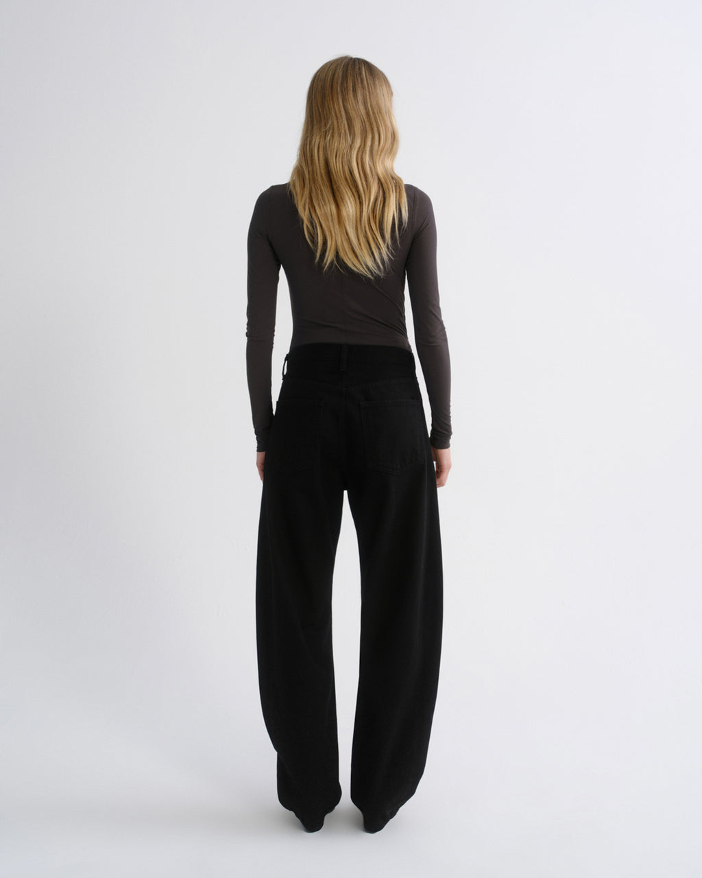A woman with long blonde hair is shown from behind wearing a dark charcoal long-sleeve top and black wide-leg trousers against a light gray studio background.