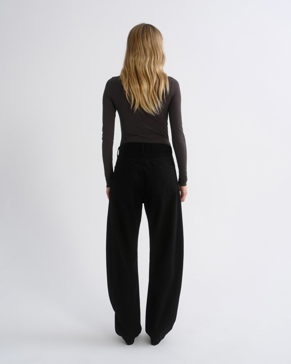 A woman with long blonde hair is shown from behind wearing a dark charcoal long-sleeve top and black wide-leg trousers against a light gray studio background.