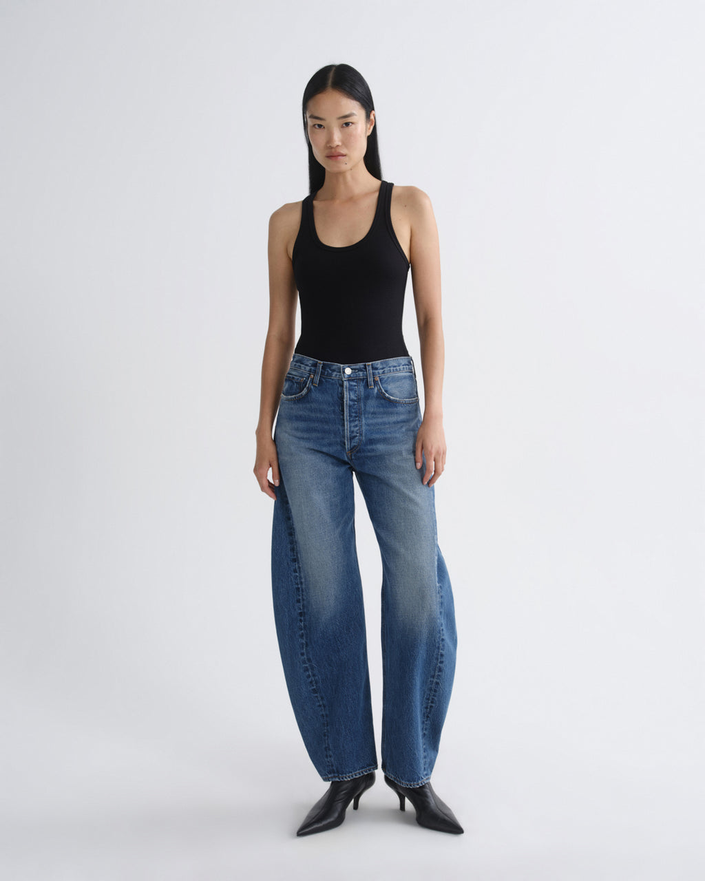 A model stands against a light gray studio background wearing a black ribbed tank top, wide-leg blue jeans, and black pointed-toe heels.