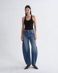 A model stands against a light gray studio background wearing a black ribbed tank top, wide-leg blue jeans, and black pointed-toe heels.