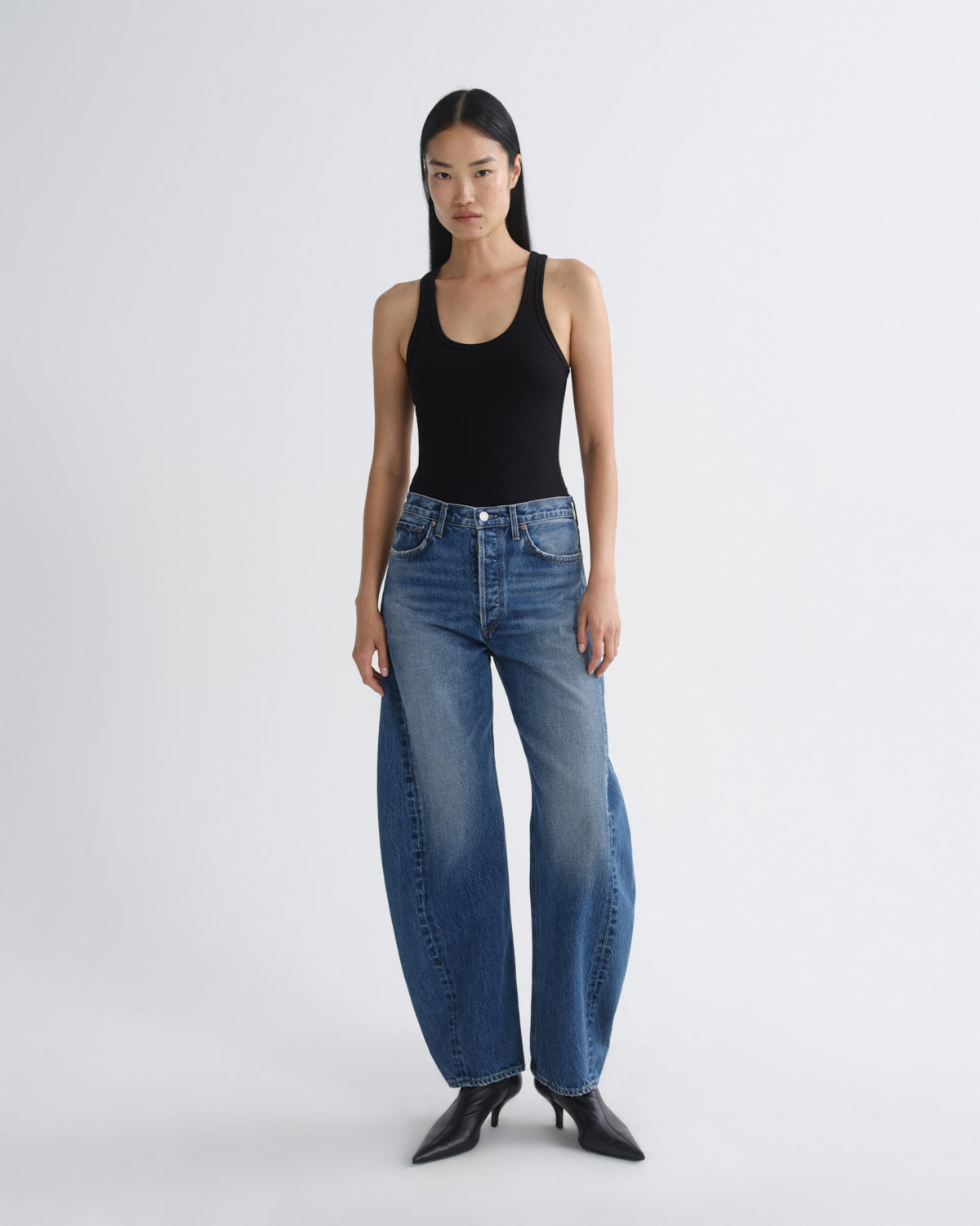 A model stands against a light gray studio background wearing a black ribbed tank top, wide-leg blue jeans, and black pointed-toe heels.
