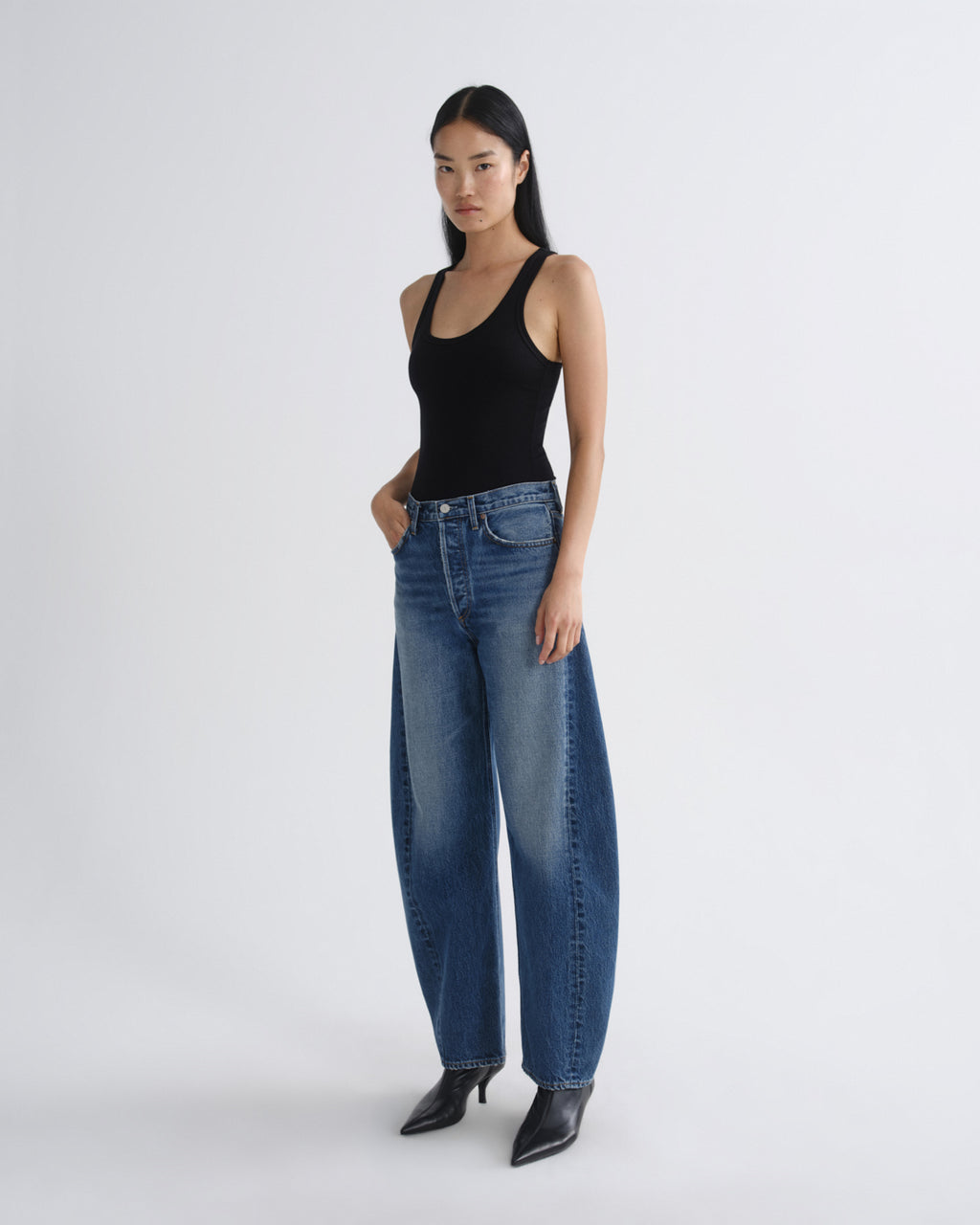 A model stands against a light gray studio background wearing a black ribbed tank top, wide-leg blue denim jeans, and black pointed-toe heeled boots.