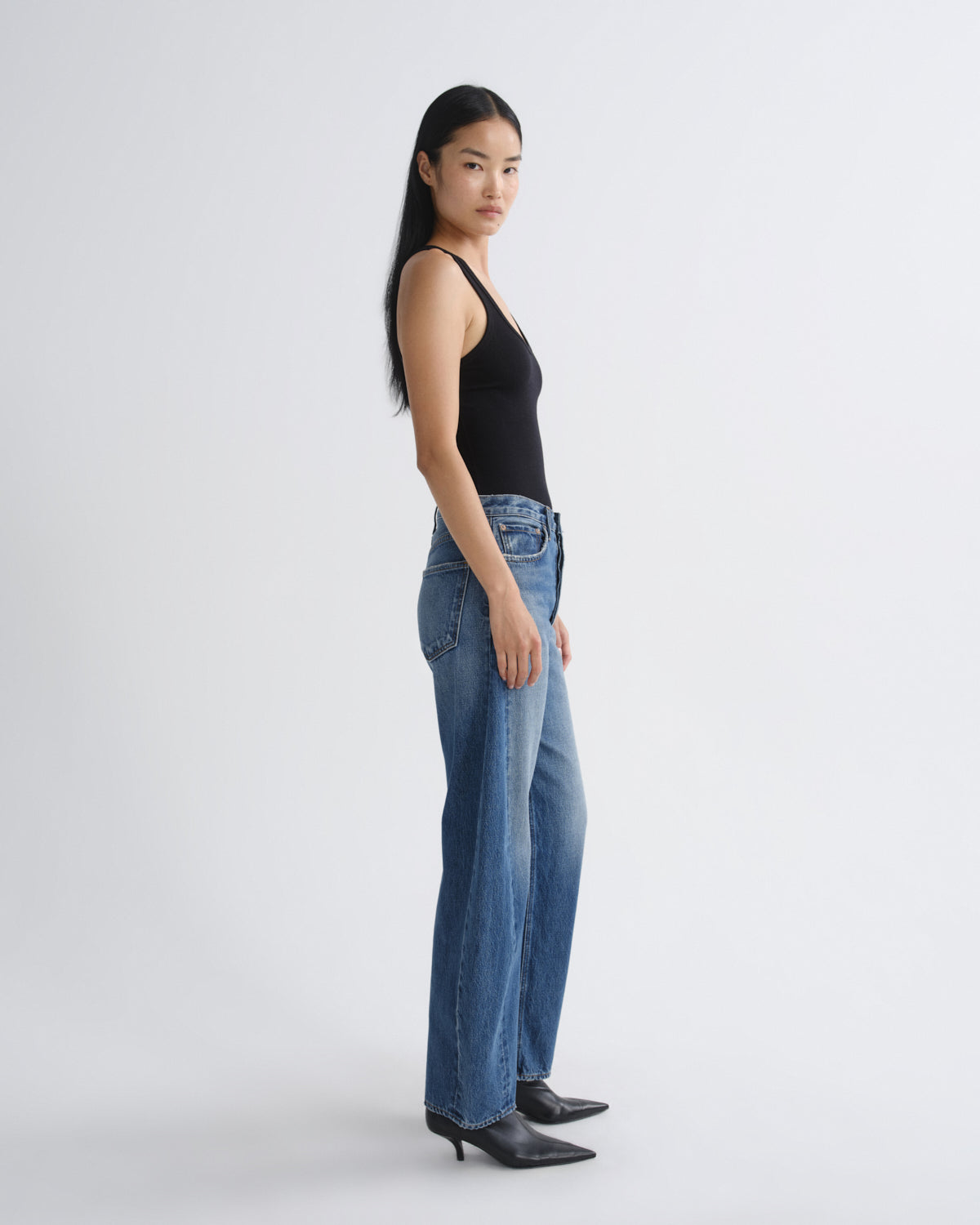 A model stands in profile wearing a black sleeveless top, blue wide-leg jeans, and black pointed-toe ankle boots against a plain light gray studio background.