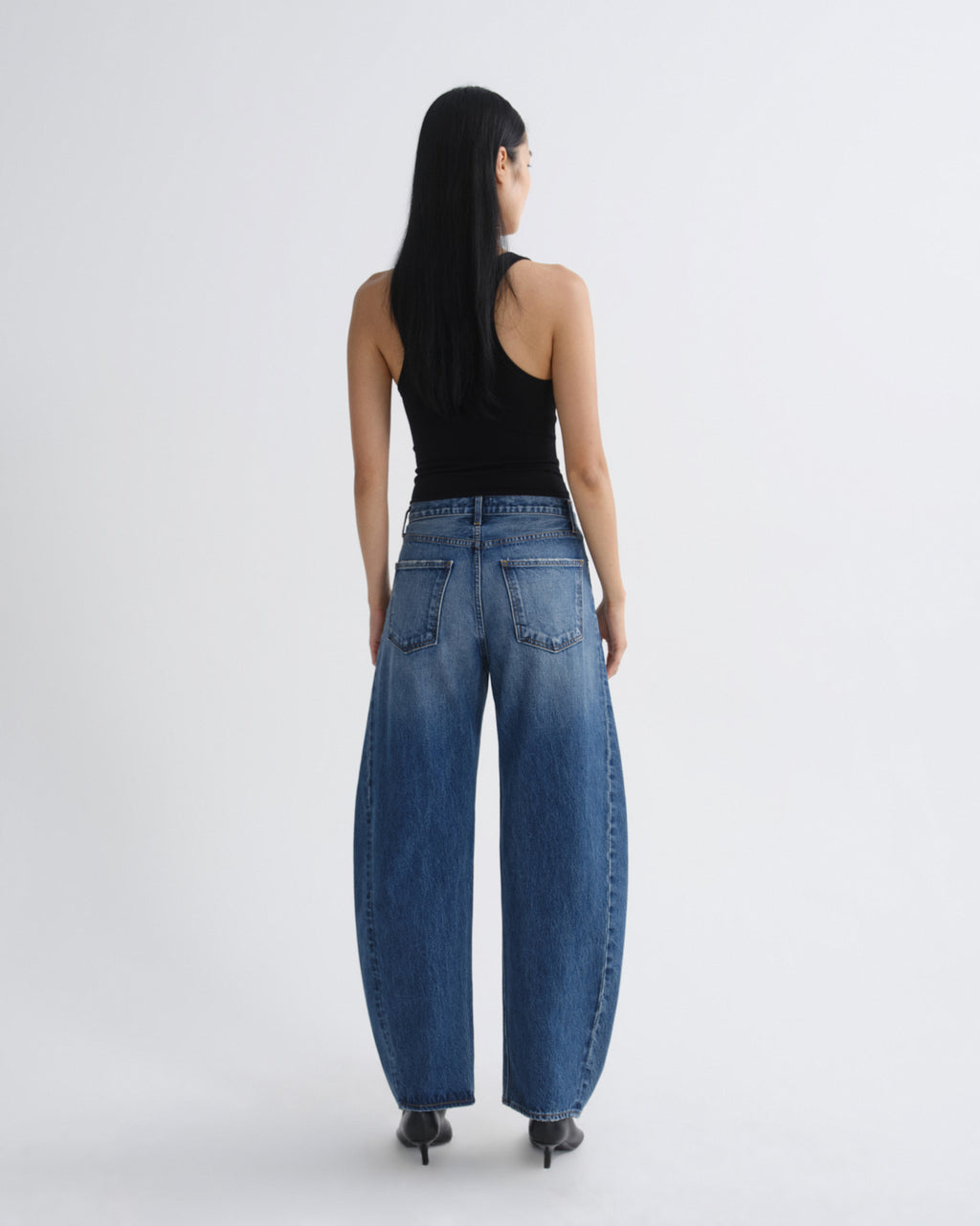 A woman viewed from behind wearing wide-leg blue denim jeans, a black tank top, and black heels against a plain light gray studio background.