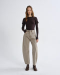 A woman stands against a light gray studio background wearing a dark brown long-sleeve top, beige wide-leg cropped pants, and dark brown ankle boots.