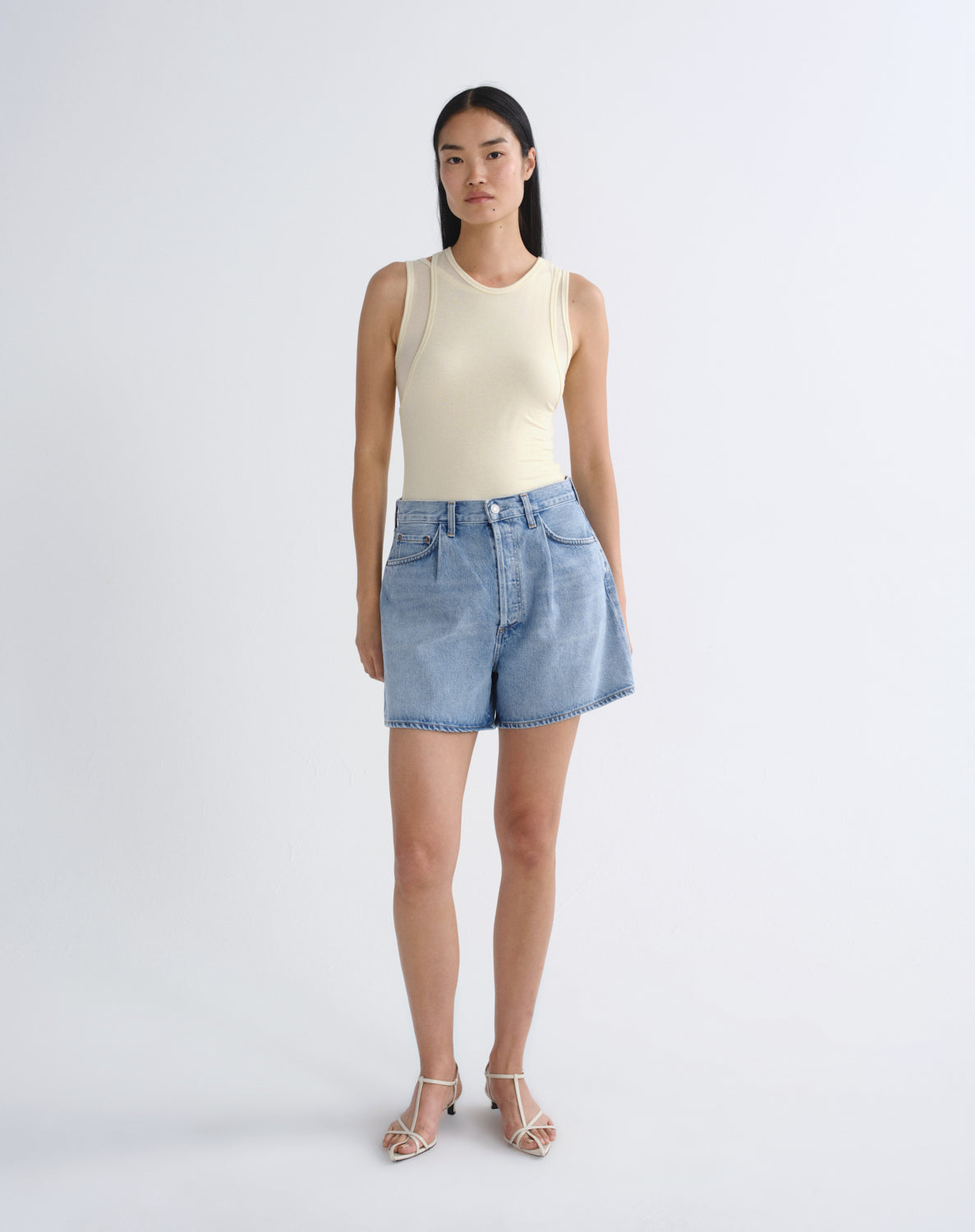 A model stands against a light gray studio background wearing a cream sleeveless top, light-wash blue denim shorts, and clear-strap high-heeled sandals.
