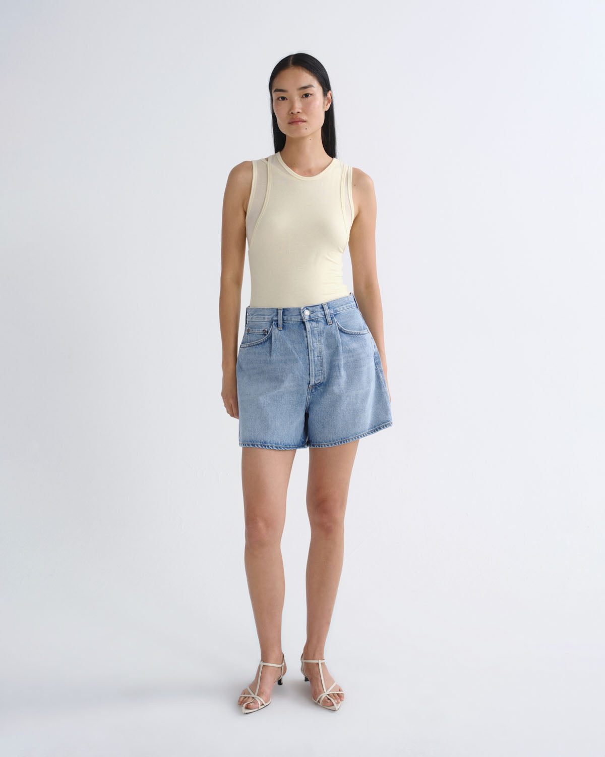 A model stands against a light gray studio background wearing a cream sleeveless ribbed tank top, light-wash blue denim shorts, and clear-strap heeled sandals.
