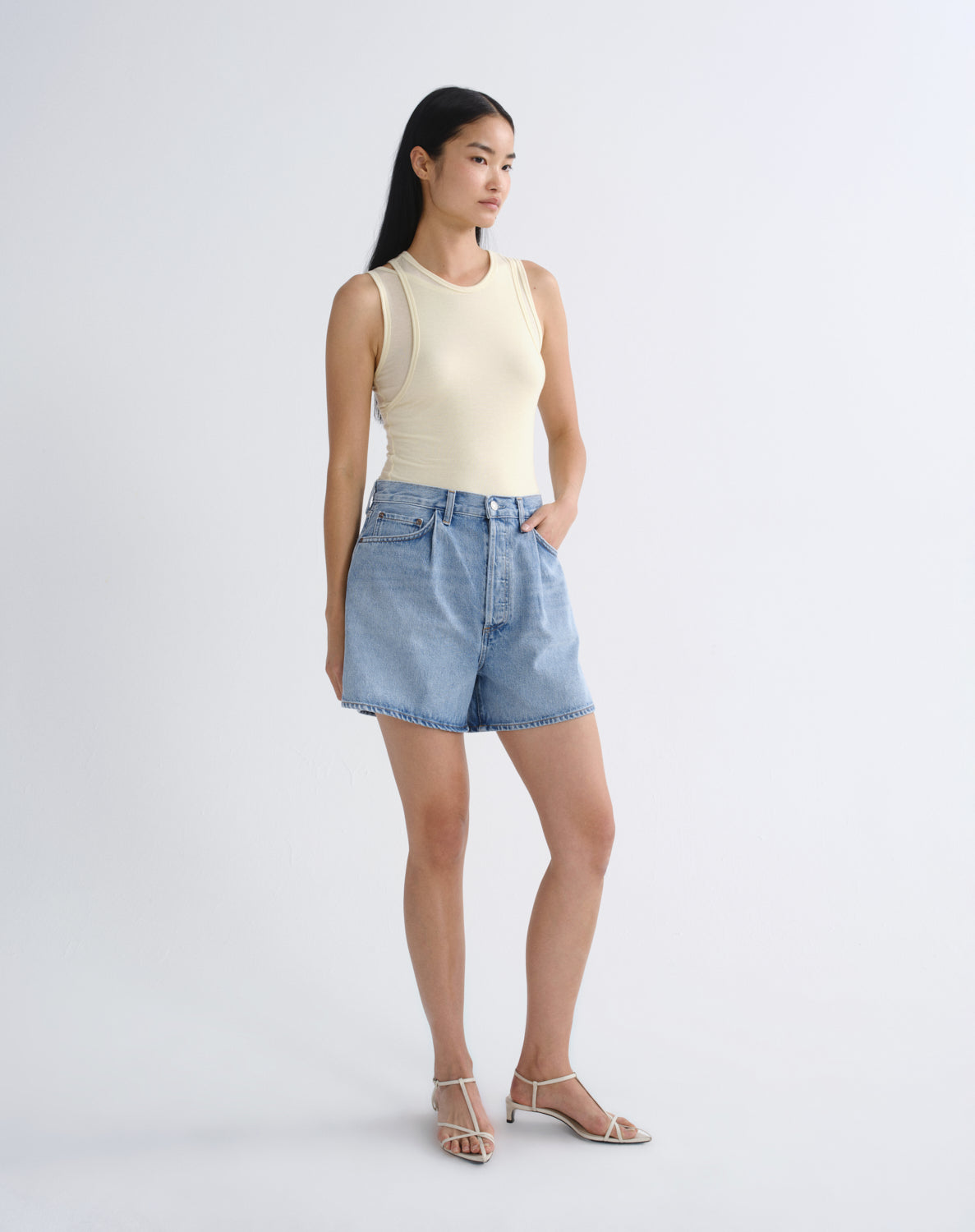A woman modeling a cream ribbed sleeveless crop top and light-wash blue denim shorts against a plain light gray studio background.