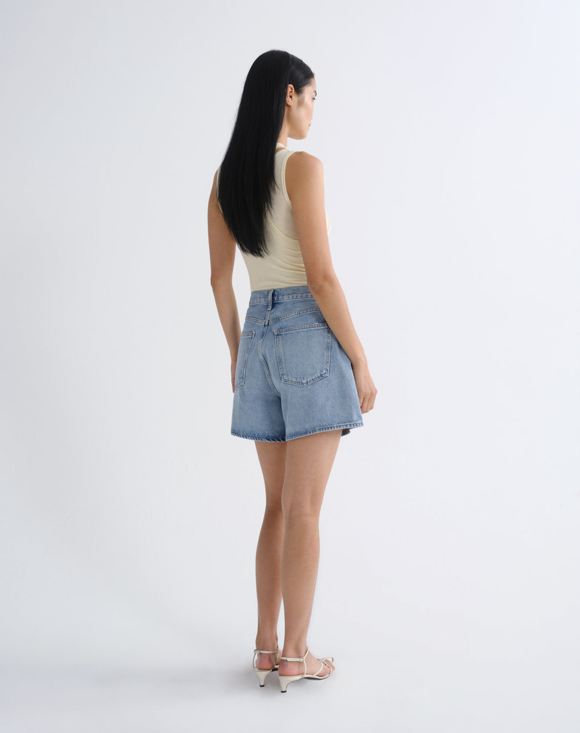 A woman is shown from the back wearing light-wash denim shorts, a light beige tank top, and light pink heeled sandals against a plain white studio background.