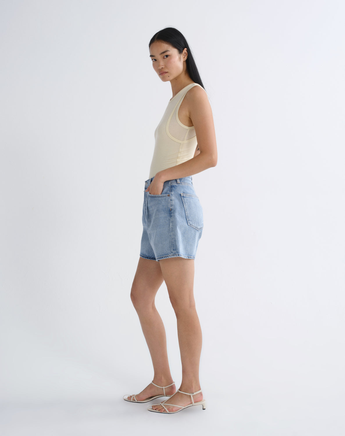 A woman stands in profile wearing a light beige ribbed tank top, light-wash denim shorts, and white strappy high-heel sandals against a plain white studio background.
